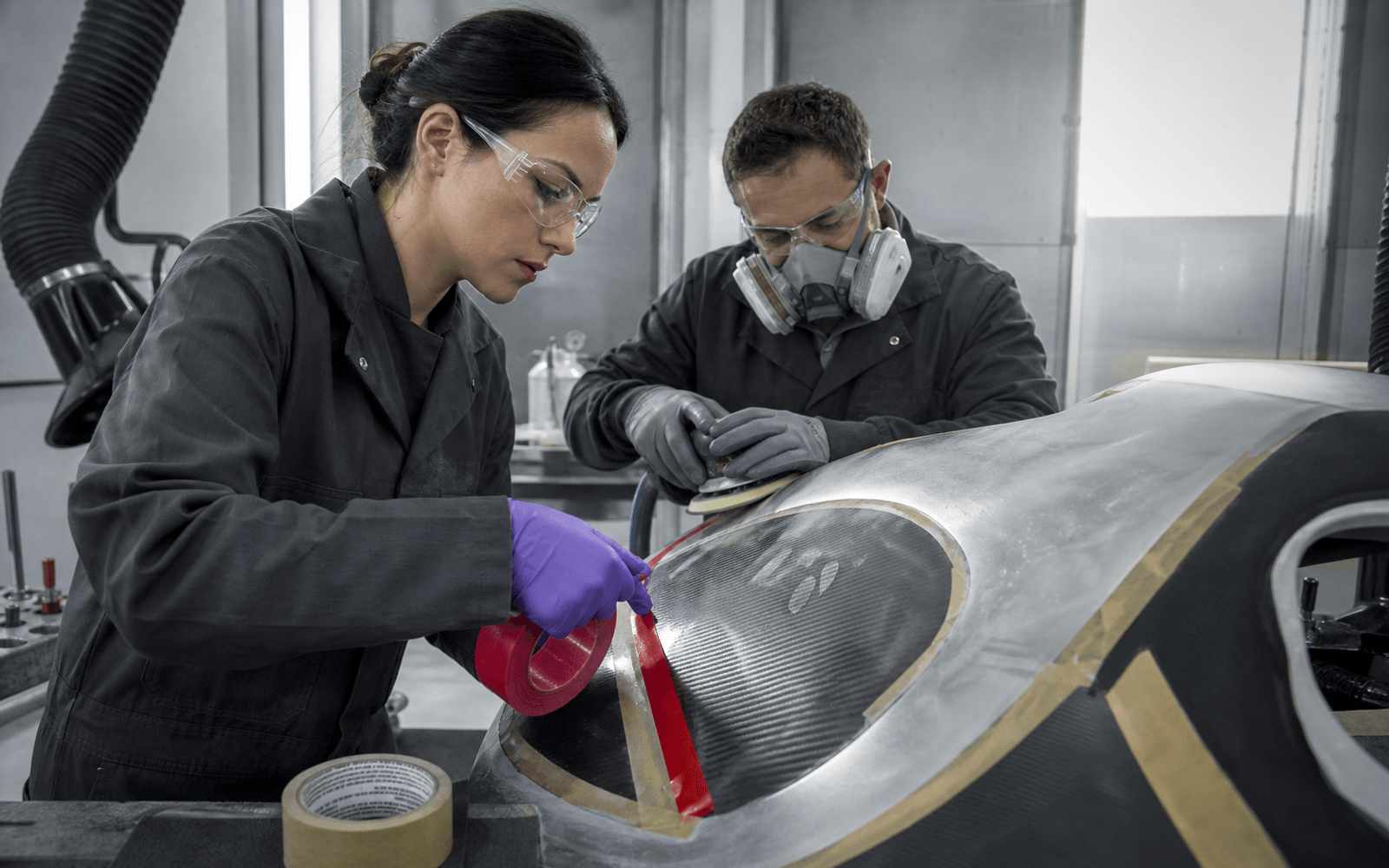 Two materials technicians apply and finish bonding tape on a carbon-fiber vehicle panel in an industrial workshop without visible branding.