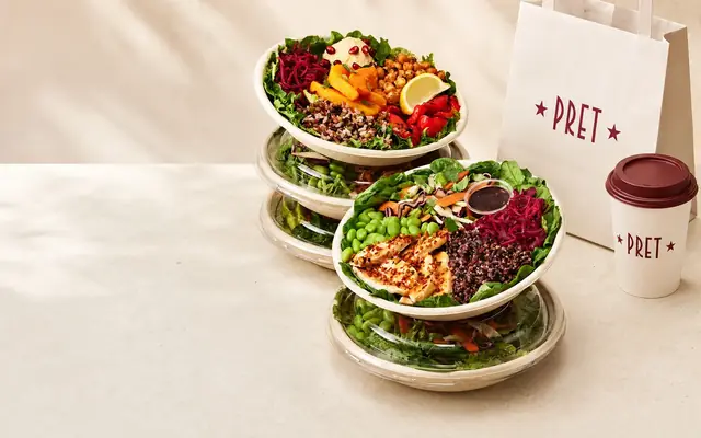 Pret A Manger salad bowls beside a branded PRET bag and takeaway cup on a clean cream counter.