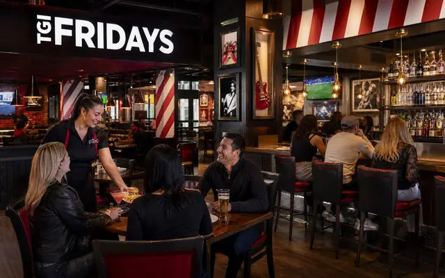 A lively active TGI Fridays bar-and-grill with guests, staff, current striped identity, and realistic restaurant proportions.